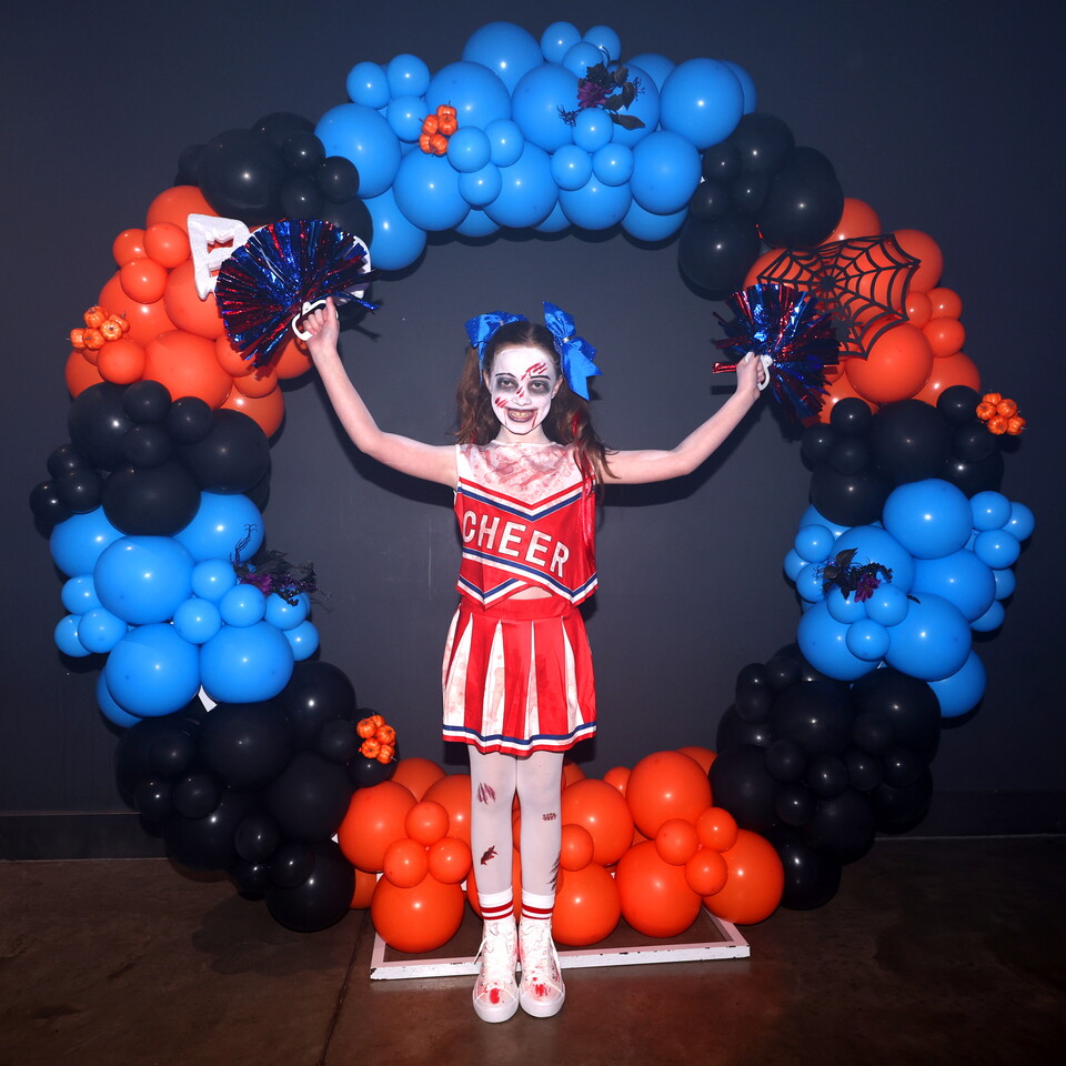 A young girl dressed as a cheerleader stands with her arms raised in front of a balloon arch.