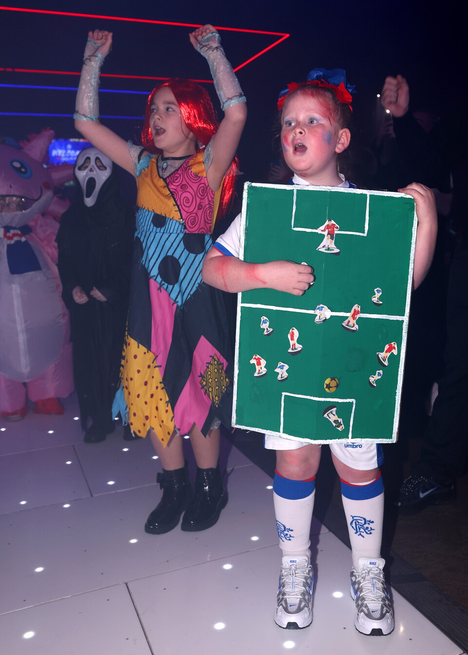 Two children on a dance floor, one wearing a Halloween costume, the other holding a football pitch sign