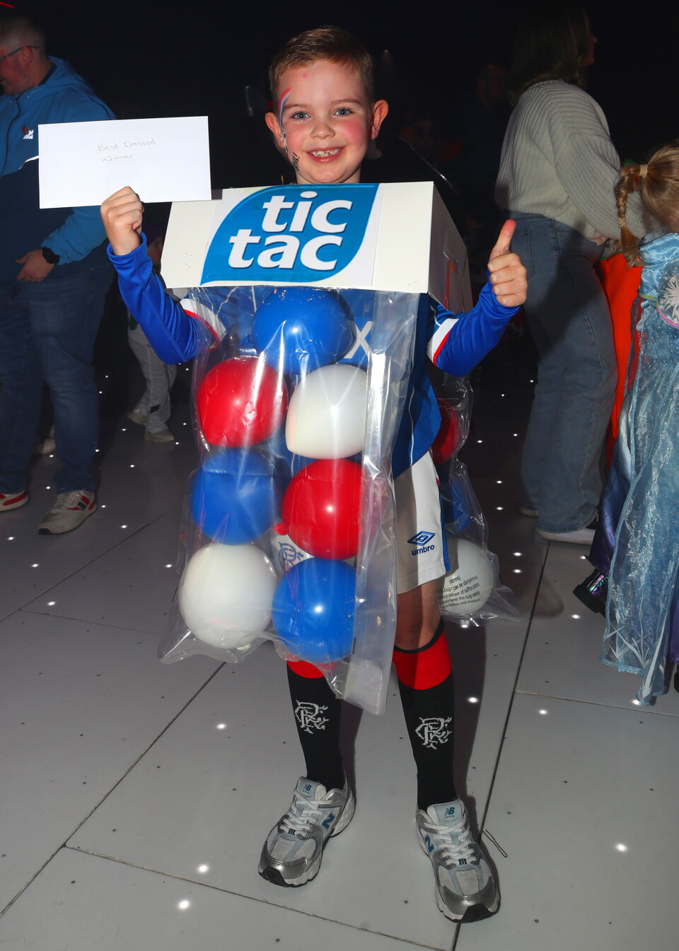 A young boy dressed in a football kit with a Tic Tac box full of balloons.
