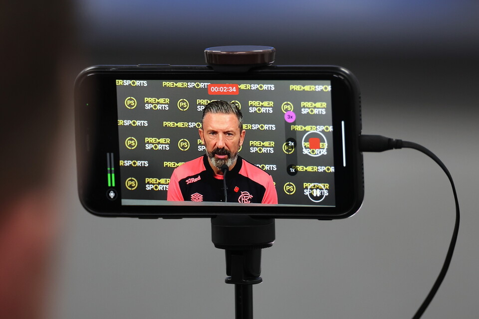 A phone displaying a man speaking into a microphone with a red and black sports team logo.