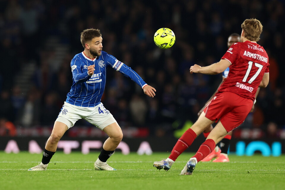Two football players, one in a blue shirt and one in a red shirt, are playing on a field with the ball in the air.