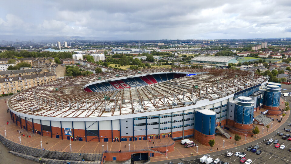Aerial view of a football stadium with red and blue trims, surrounded by parked cars and trees.