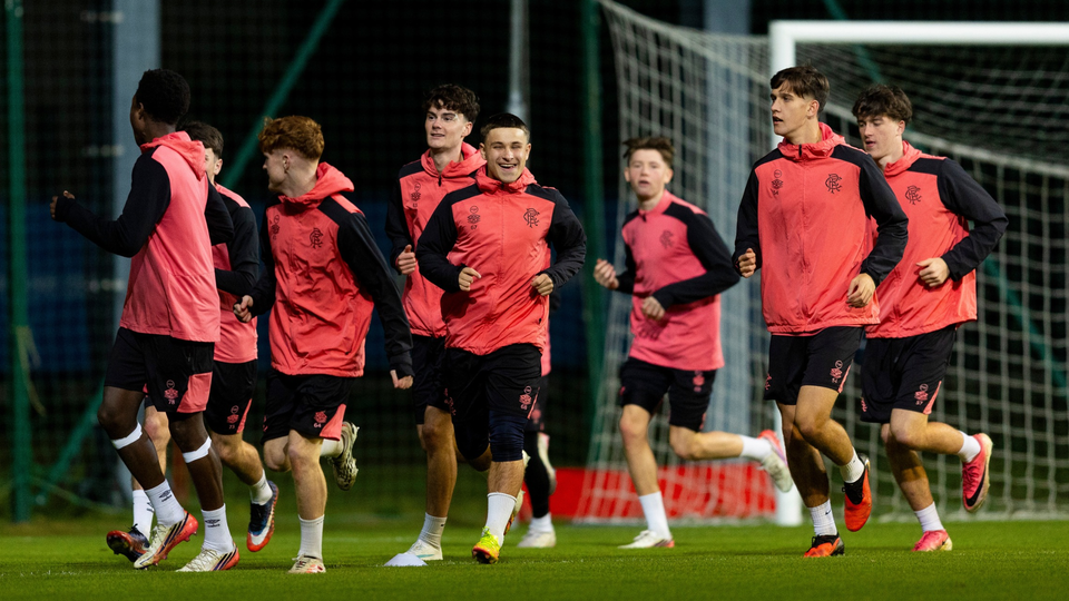 Seven players in red shirts and black shorts running towards a goalpost on a football pitch.