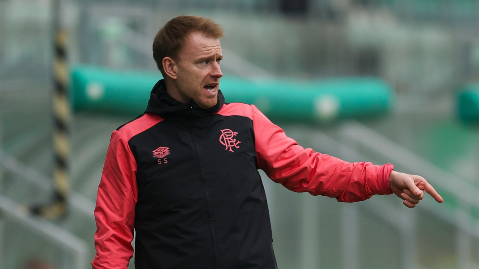 Football coach with red and black top directs players on a pitch