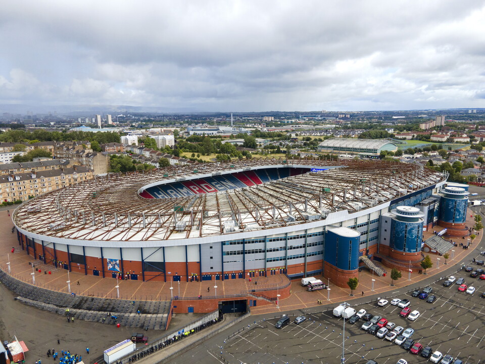 Aerial view of a football stadium with red and blue trims, surrounded by parked cars and trees.