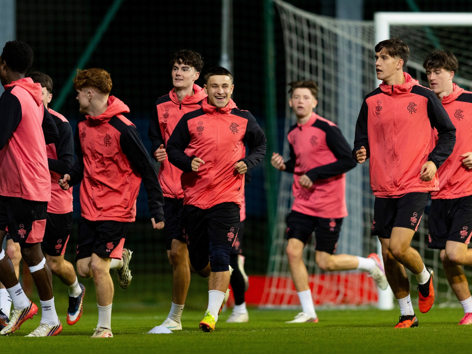 Seven players in red shirts and black shorts running towards a goalpost on a football pitch.