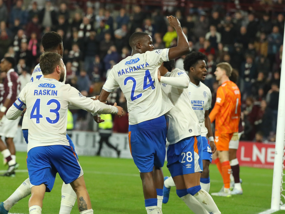 Players celebrate a goal in a football match, wearing white and blue kits with numbers 24, 43, and 18.